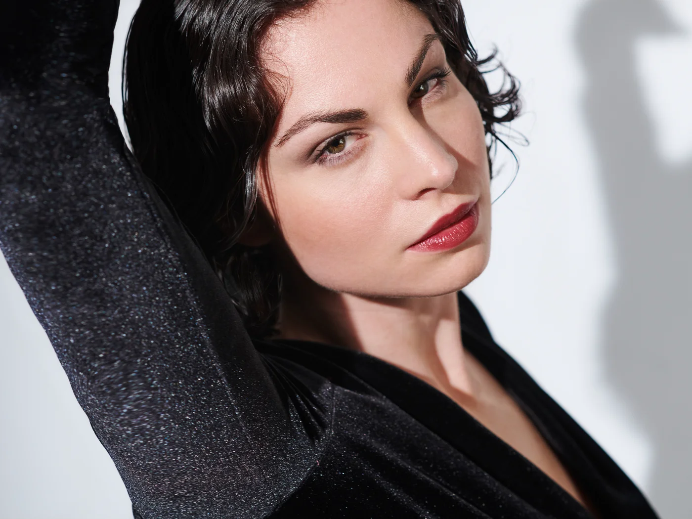 Die Schlossevents Frau mit rotem Lippenstift und schwarzem Oberteil hebt Arm vor weißem Hintergrund