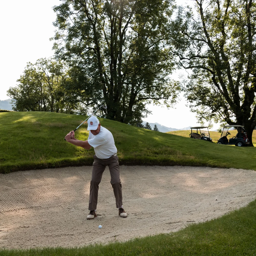 Ein ganzes Jahr in Bewegung Mann schlägt Golfball aus Bunker auf sonnigem Golfplatz mit zwei Golfwagen im Hintergrund