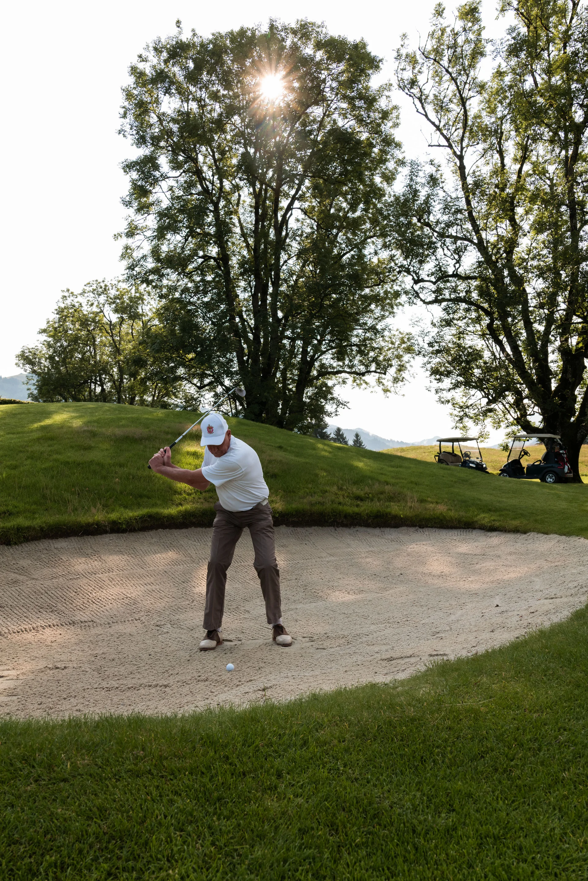 Das Golfhotel in Kitzbühel Mann schlägt Golfball aus Bunker auf sonnigem Golfplatz mit zwei Golfwagen im Hintergrund