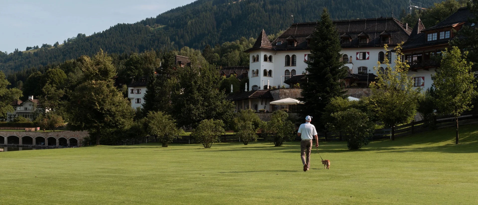 Ins Hotel mit Hund in Kitzbühel Mann geht mit Hund auf einer grünen Wiese vor einem bergigen Schloss