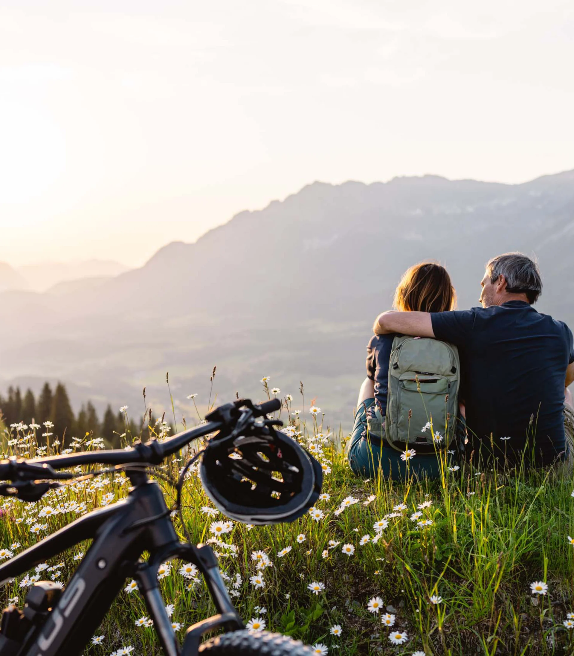 Ein ganzes Jahr in Bewegung Paar sitzt auf einer Blumenwiese, blickt auf Berge bei Sonnenuntergang
