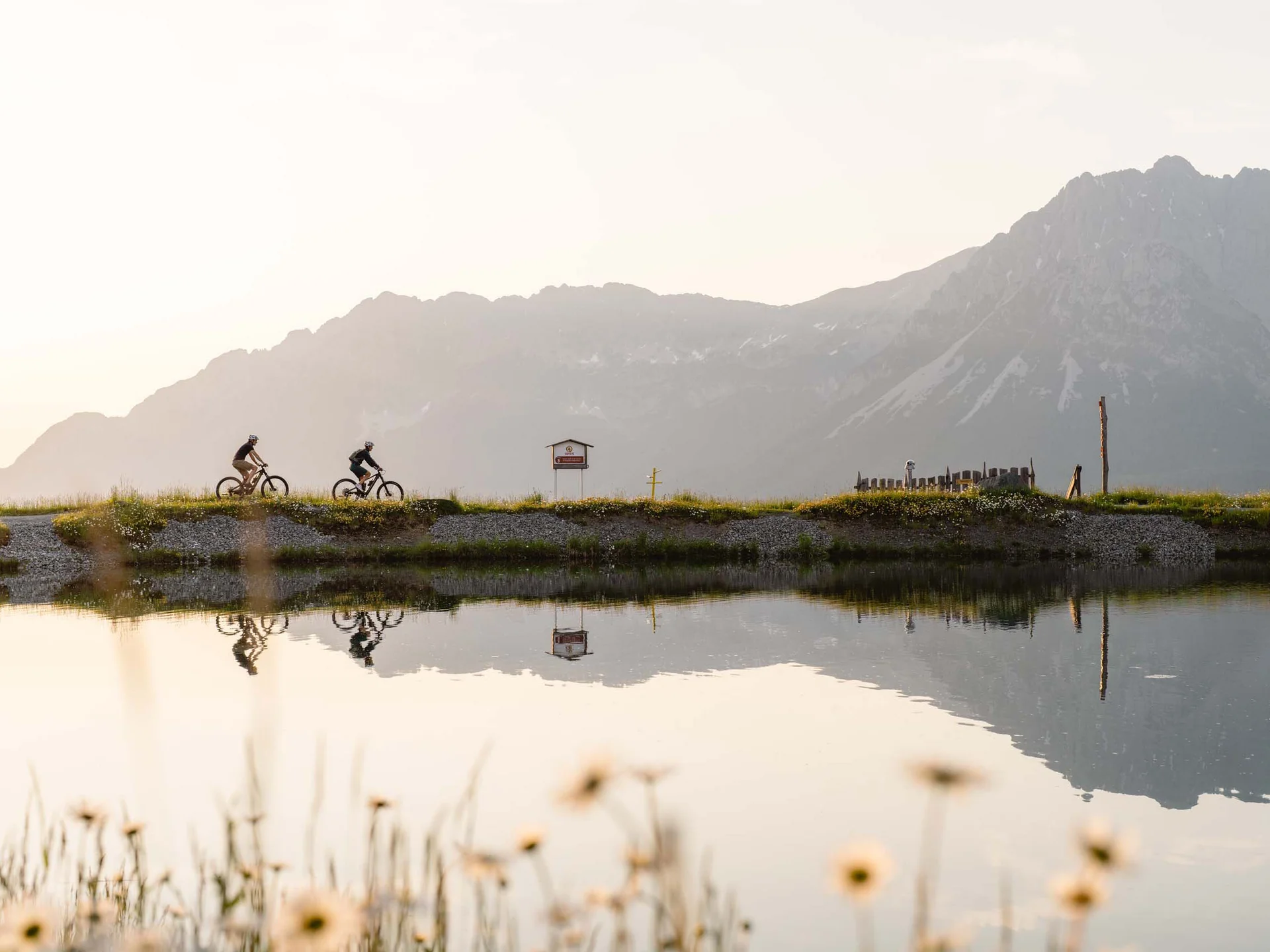 Bilder, die Sie sehen müssen. Zwei Radfahrer fahren entlang eines Sees mit Bergen im Hintergrund bei Sonnenuntergang