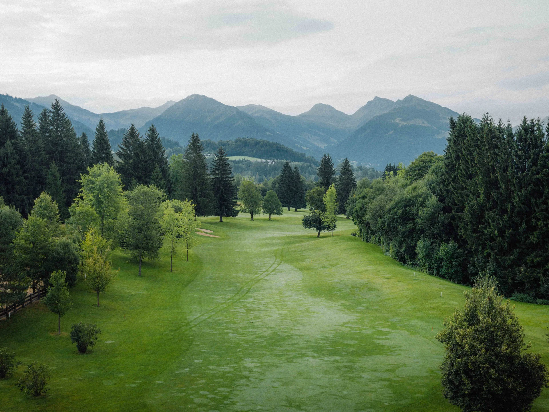 Bilder, die Sie sehen müssen. Grüner Golfplatz mit Bäumen und Bergen im Hintergrund unter bewölktem Himmel
