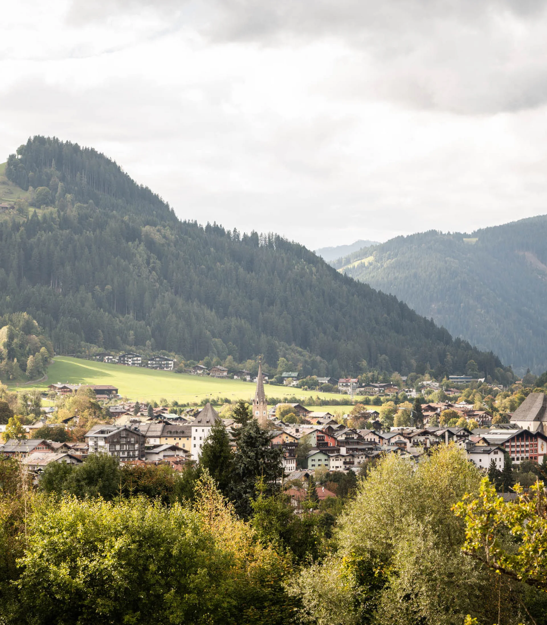 Mit der Familie ins Hotel? Tirol ist die richtige Wahl. Bergdorf umgeben von Wäldern und Bergen unter bewölktem Himmel