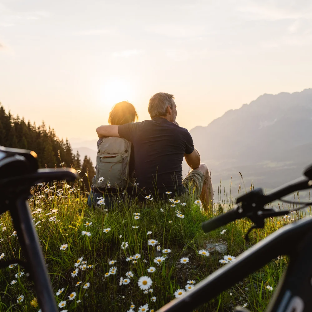 Ein ganzes Jahr in Bewegung Paar sitzt auf einer Wiese mit Blumen und blickt auf Berge im Sonnenuntergang