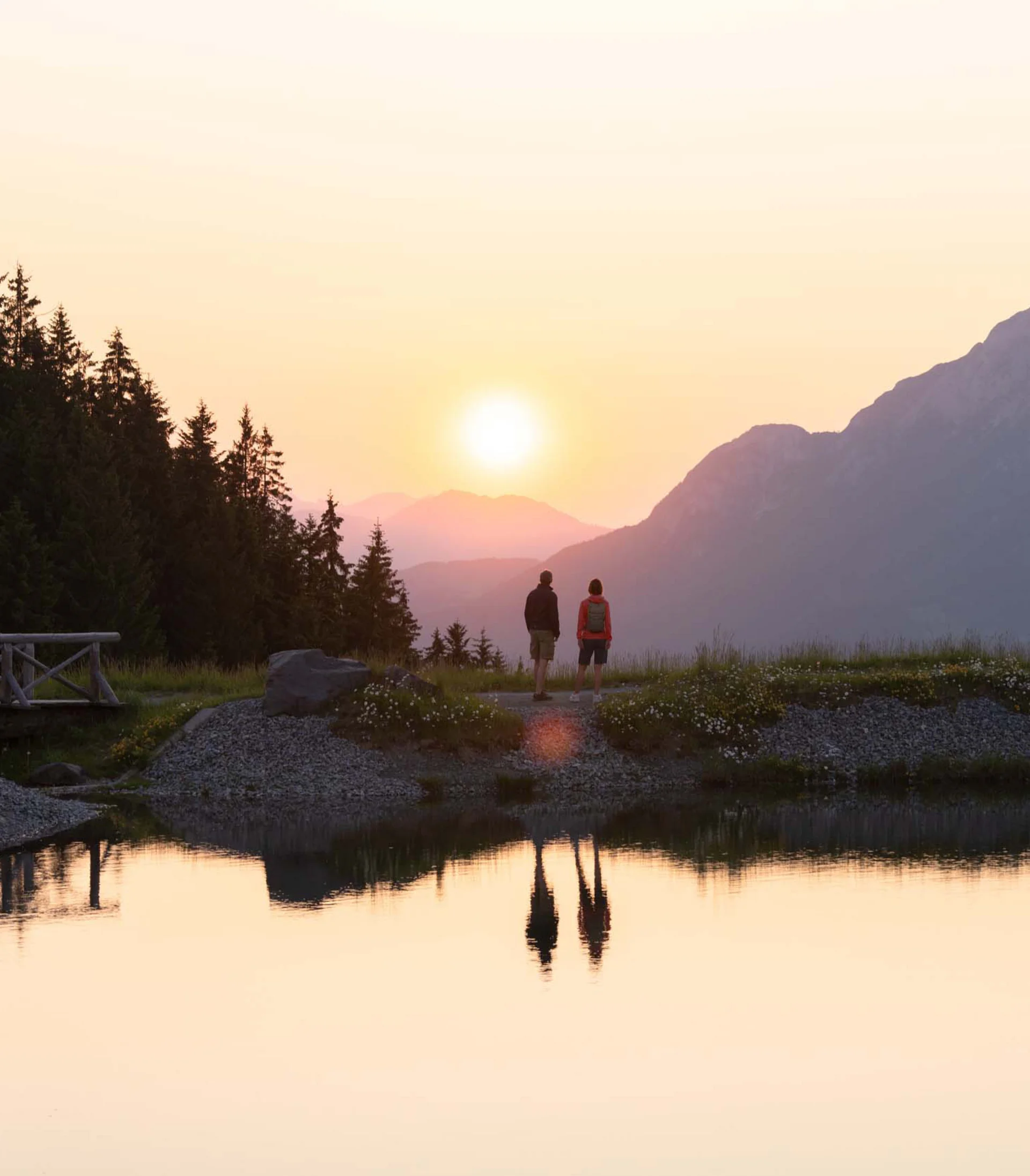 Rund ums Bikehotel in Kitzbühel wandern Zwei Personen betrachten den Sonnenuntergang an einem Bergsee mit Wald und Brücke