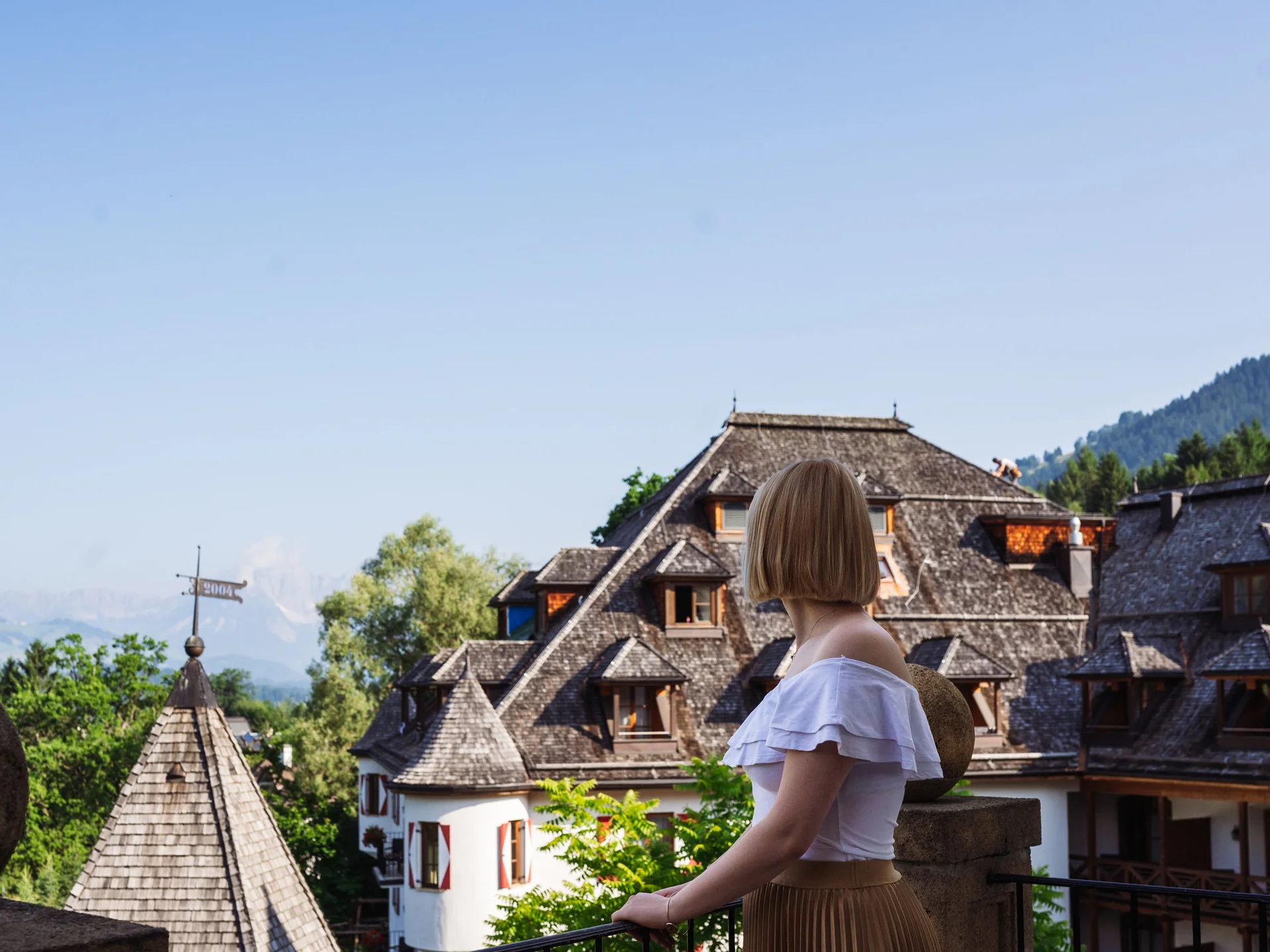Bilder, die Sie sehen müssen. Frau schaut von Balkon auf traditionelles Haus mit Bergkulisse bei klarem Himmel