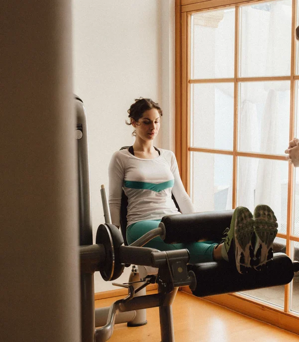 Fitness und Yoga im Hotel in Tirol Frau trainiert Beinmuskulatur an Maschine, Mann unterstützt als Trainer