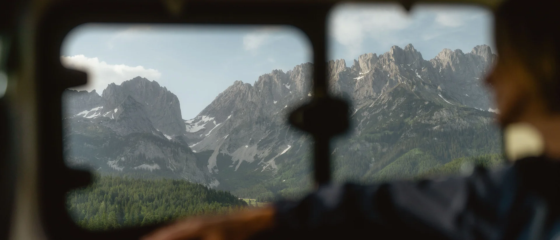 Wonach steht Ihnen heute der Sinn in Kitzbühel? Berglandschaft mit Nadelwald durch ein Busfenster gesehen, Person unscharf im Vordergrund