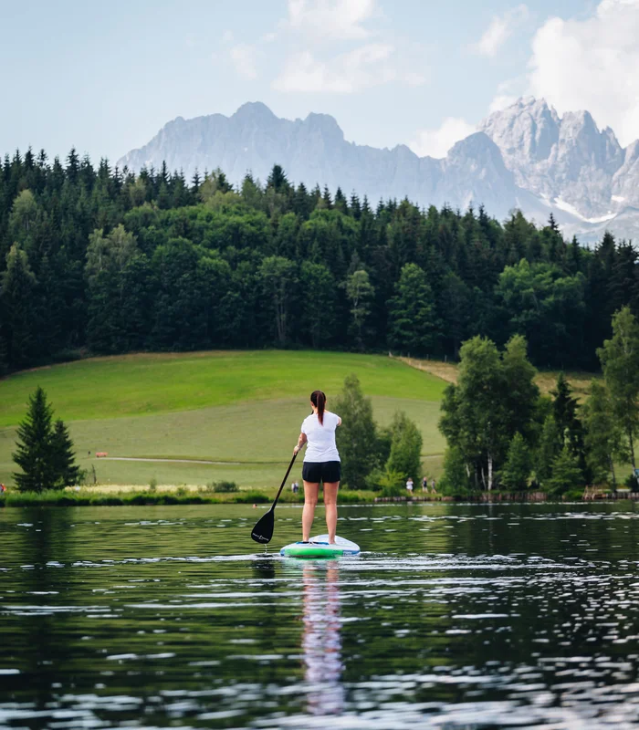 Wonach steht Ihnen heute der Sinn in Kitzbühel? Eine Frau paddelt auf einem See mit Bergen und Wald im Hintergrund
