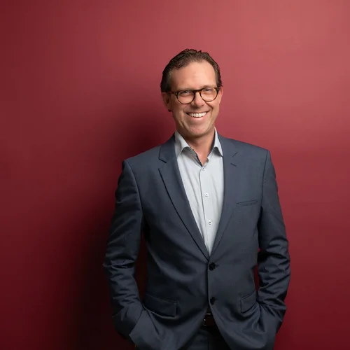 Restaurant Kitzbühel | Genuss im Schlosshotel Lächelnder Mann in Brille und Anzug vor rotem Hintergrund