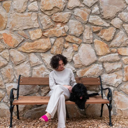 Ins Hotel mit Hund in Kitzbühel Frau sitzt auf Parkbank und streichelt schwarzen Hund vor Steinmauer