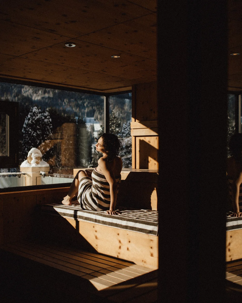 In Kitzbühel ein Hotel mit Pool, bitte. Frau sitzt in Sauna mit Handtuch und blickt auf winterliche Landschaft