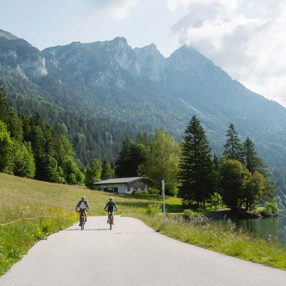 Ein ganzes Jahr in Bewegung Zwei Radfahrer fahren auf Straße neben See mit Bergen im Hintergrund