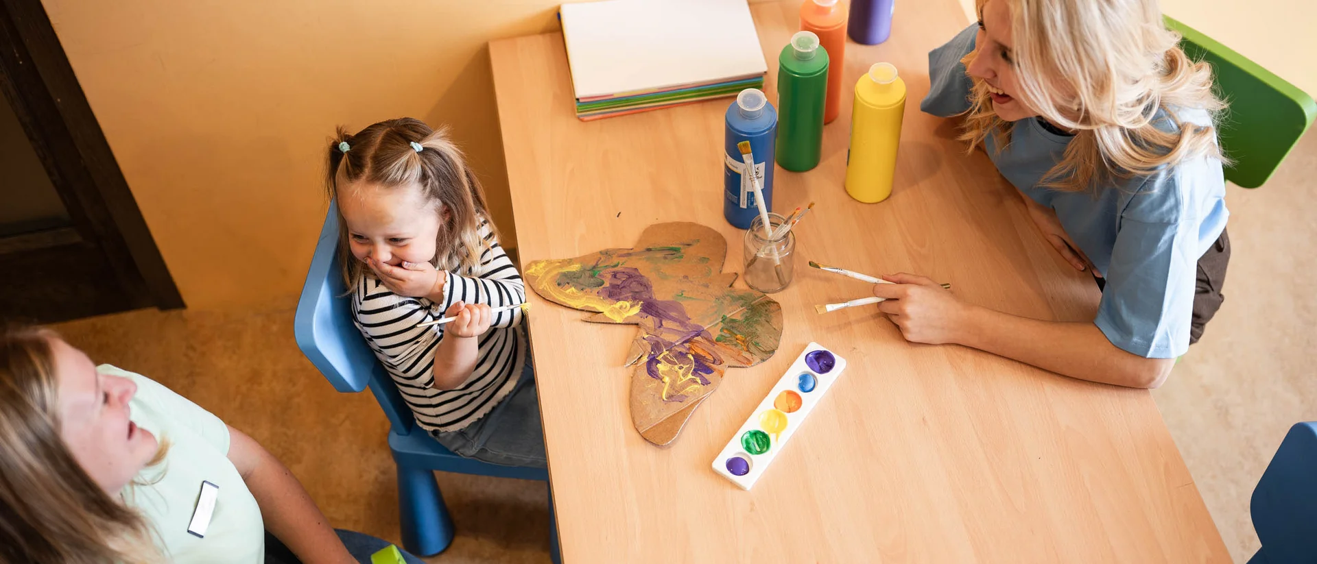 Mit der Familie ins Hotel? Tirol ist die richtige Wahl. Kinder und Betreuerinnen lachen zusammen beim Malen am Tisch