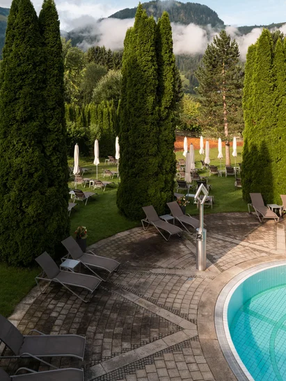 In Kitzbühel ein Hotel mit Pool, bitte. Poolbereich mit Liegestühlen und grünen Bäumen vor Bergen unter bewölktem Himmel